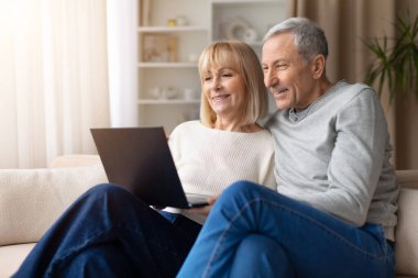 Üst düzey çift evde dizüstü bilgisayar kullanırken kanepede dinleniyorlar. Olgun erkek ve kadın internette geziniyor veya dijital yaşam tarzı ve modern teknoloji kullanımını temsil eden çevrimiçi içeriği izliyor