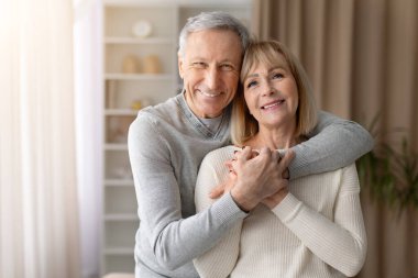 Evdeki oturma odasında yan yana dururken kameraya sarılıp gülümseyen mutlu yaşlı çift. Aşkı, aile bağını, güveni ve uzun vadeli ilişkiyi temsil eden olgun bir adam.
