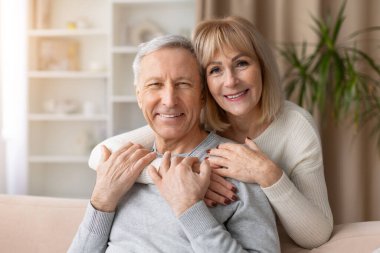 Evde kanepede kucaklaşan gülümseyen yaşlı çiftin portresi. Olgun beyaz erkek ve kadın kameraya bakıyor. İlişkiyi, aile mutluluğunu ve rahat emekliliği temsil ediyor.