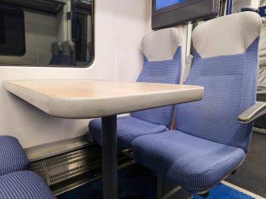 Boş bir banliyö vagonunda, içi boş koltuklar ve masalarla dolu bir iç mekan.