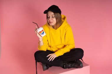 Sarı kapüşonlu ceketli, gökkuşağı kalpli bir kupa tutan sırıtan genç bir LGBTQ kadını..