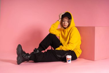 Kapüşonlu ve siyah şapkalı bir kadın kağıt bardağın yanında aşk ve gökkuşağı renkli bir kalp yazar.