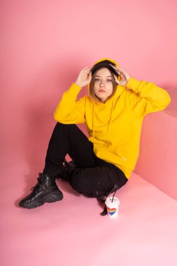 Sarı kapüşonlu ve şapkalı üzgün genç bir LGBT kadın güneşliği tutuyor ve objektife bakıyor, yanında gökkuşağı kalpli bir kupa var ve yazıda aşk yazıyor.