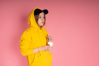 Sarı kapüşonlu ceket giyen, pembe arka planda gökkuşağı kalpli bir kupa tutan genç LGBTQ kadını.