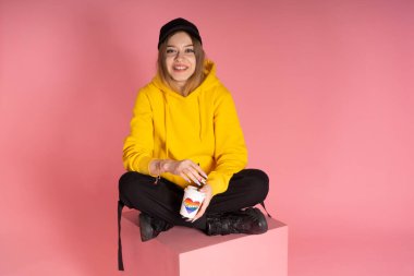 Sarı kapüşonlu ceketli, gökkuşağı kalpli bir kupa tutan sırıtan genç bir LGBTQ kadını..