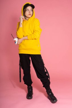 Sarı kapüşonlu ceket giyen, pembe arka planda gökkuşağı kalpli bir kupa tutan genç LGBTQ kadını.