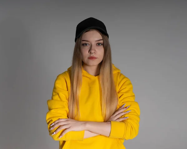 in the studio, a girl in a yellow sweatshirt and a black cap looks into the lens, the background behind her is gray