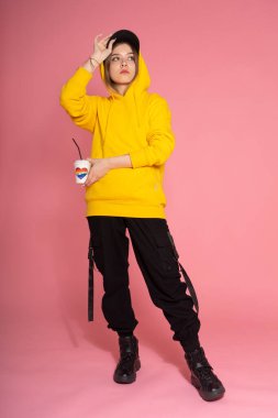 Genç LGBTQ üzgün kadın sarı kapüşonlu ceket giyiyor. Elinde pembe arka planda, gökkuşağı kalpli bir bardak tutuyor.