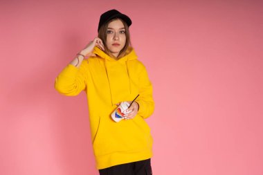 Sarı kapüşonlu ceket giyen, pembe arka planda gökkuşağı kalpli bir kupa tutan genç LGBTQ kadını.