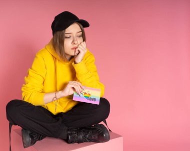 Sarı kapüşonlu ceket giyen üzgün bir kadın ve elinde LGBTQ gökkuşağı aşk kartı tutan pembe arka planda üzgün ya da melankolik görünen bir şapka..