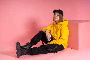 Sarı kapüşonlu ceketli, gökkuşağı kalpli bir kupa tutan sırıtan genç bir LGBTQ kadını..