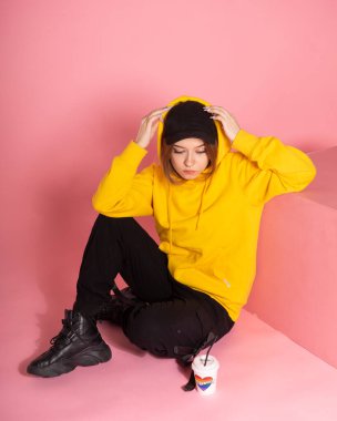 Genç LGBTQ üzgün kadın sarı kapüşonlu ceket giyiyor. Elinde pembe arka planda, gökkuşağı kalpli bir bardak tutuyor.