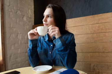 Mavi kot gömlekli bir kız bir kafede oturur ve pencereden dışarı bakar, elinde yeşil çay olan beyaz bir kupayla.