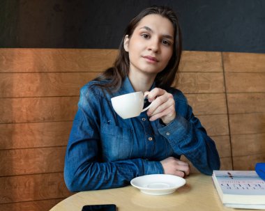 Mavi kot gömlekli bir kız bir kafede oturuyor ve elinde bir kupa yeşil çayla bir lense bakıyor.