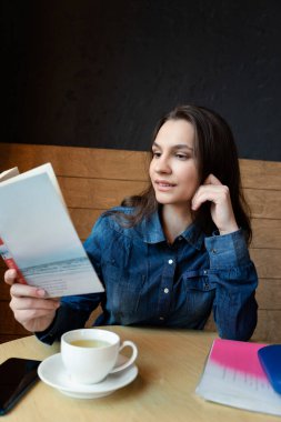 Mavi kot gömlekli düşünceli bir kız bir kafede oturur ve bir kitap okur. Masada beyaz bir fincanda yeşil çay vardır.