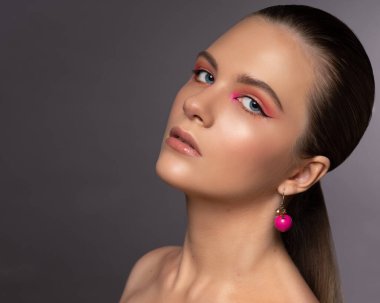 Mavi gözlü, yuvarlak pembe küpeli ve uzun kahverengi saçlı harika profesyonel makyajlı güzel bir genç kızın yüksek güzellikte fotoğrafı. Gri arka planda poz veriyor. Yakın plan. Stüdyo çekimi. Portre