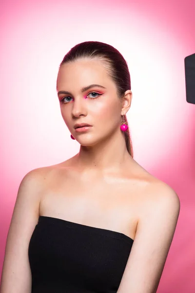Mavi gözlü, yuvarlak pembe küpeli ve uzun kahverengi saçlı harika profesyonel makyajlı güzel bir genç kızın yüksek güzellikte fotoğrafı. Pembe arka planda poz vermek. Yakın plan. Stüdyo çekimi. Portre