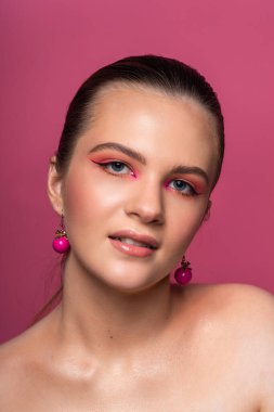 Mavi gözlü, yuvarlak pembe küpeli ve uzun kahverengi saçlı harika profesyonel makyajlı güzel bir genç kızın yüksek güzellikte fotoğrafı. Pembe arka planda poz vermek. Yakın plan. Stüdyo çekimi. Portre
