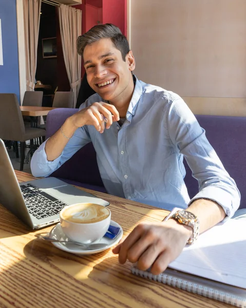 Neşeli genç bir iş adamı iş arkadaşlarıyla dizüstü bilgisayarda iletişim kuruyor, güzel bir kafede, kapuçino ve masanın üzerinde bir defter var. Uzak çalışma.