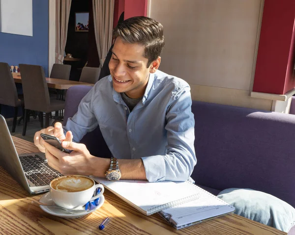 Neşeli öğrenci telefonla video görüşmesiyle iletişim kuruyor. Kafede bir adam. Masanın üstünde iki not defteri vardı. Elinde beyaz bir bardak kapuçino tutuyordu..