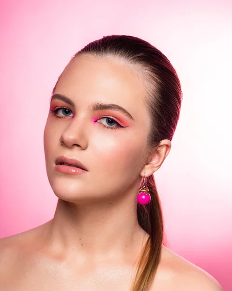 High beauty photo of a lovely young girl with blue eyes, round pink earrings and wonderful professional makeup, with long brown hair. Posing over rosy background. Close-up. Studio shot. Portrait