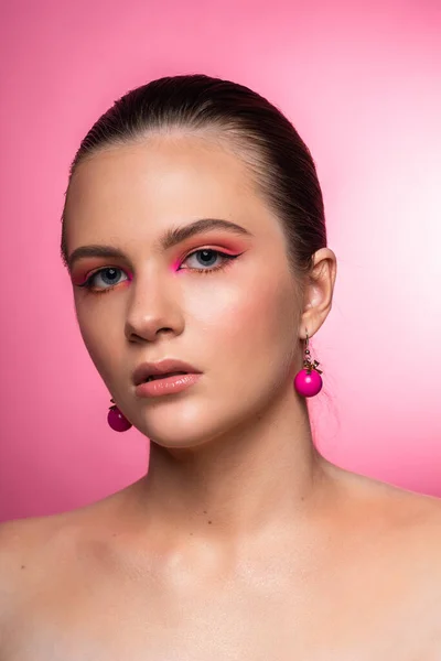 Mavi gözlü, yuvarlak pembe küpeli ve uzun kahverengi saçlı harika profesyonel makyajlı güzel bir genç kızın yüksek güzellikte fotoğrafı. Pembe arka planda poz vermek. Yakın plan. Stüdyo çekimi. Portre