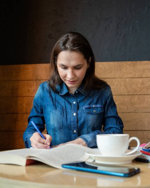 Kutsanmış bir kız ahşap bir cetvel kullanarak mavi bir keçeli kalemle duygu takvimi çizer. Önünde bir kitap ve yeşil çaylı beyaz bir kupa vardır.