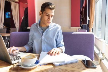Düşünceli öğrenci dişçi bir kafede oturur ve bir ders verir. El defterin üzerinde yatıyor. Masanın üzerinde bir telefon, bir laptop ve bir fincan kahve var. Karantinada uzaktan çalışma