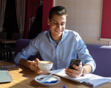 Neşeli öğrenci telefonla video görüşmesiyle iletişim kuruyor. Kafede bir adam. Masada bir dizüstü bilgisayar, iki not defteri ve bir kupa kapuçino var..