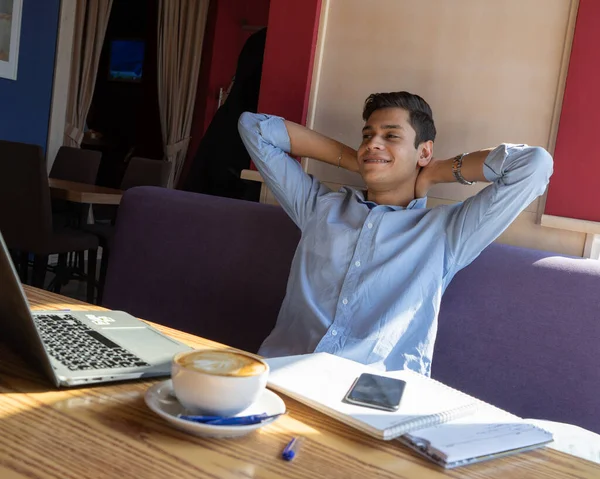 Genç serbest çalışan mutlu ve rahatlamış. Projeyi tamamladı. İş uzaktan hallediliyor, bir adam bir kafede. Masada bir dizüstü bilgisayar, iki not defteri ve bir kupa kapuçino var..