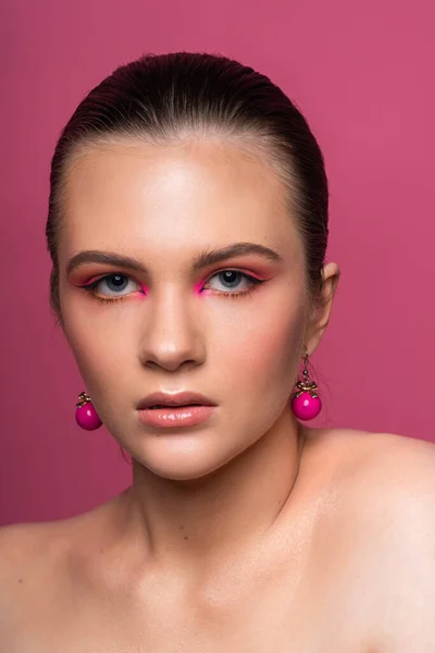 Mavi gözlü, yuvarlak pembe küpeli ve uzun kahverengi saçlı harika profesyonel makyajlı güzel bir genç kızın yüksek güzellikte fotoğrafı. Pembe arka planda poz vermek. Yakın plan. Stüdyo çekimi. Portre