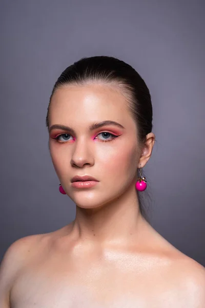 Harika profesyonel makyajlı, pembe küpeli, uzun kahverengi saçlı güzel bir genç kızın yüksek güzellikte fotoğrafı. Gri arka planda poz vermek. Yakın plan. Stüdyo çekimi. Portre