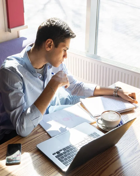 Genç bir işadamı biyoloji projesini düşünüyor. Yanında bir laptop, bir telefon ve kahve ile birlikte beyaz bir bardak var. Bir kafede.