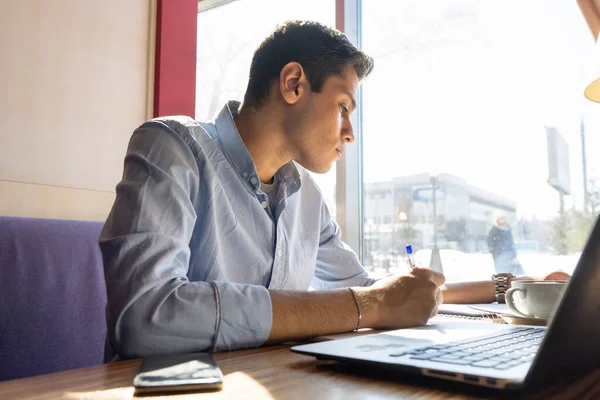 Genç bir işadamı proje için tahmin yürütür. Yanında bir dizüstü bilgisayar, bir telefon ve kahveli beyaz bir bardak vardır.