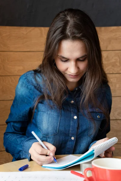 カフェに座っている青いデニムシャツの真面目な女の子はペンでメモ帳にメモを作ります — ストック写真