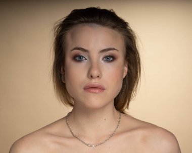 Heterokromisi olan, burun piercingli, tek kulağı tıkalı ve garip bir saç stili olan seksi bir kız. Objektife bakınca, inanılmaz bir profesyonel makyajı ve boynunda bej arka planda gümüş bir zincir var.