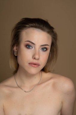 Heterokromisi olan, burun piercingli, tek kulağına takılan ve garip saç stili olan seksi bir kız. Boynunda inanılmaz bir profesyonel makyaj ve gümüş zincir var. Bej arka plan. Stüdyo çekimi