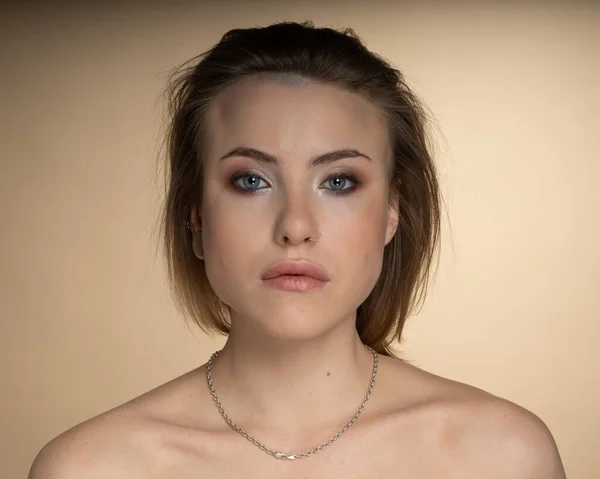 Heterokromisi olan, burun piercingli, tek kulağı tıkalı ve garip bir saç stili olan seksi bir kız. Objektife bakınca, inanılmaz bir profesyonel makyajı ve boynunda bej arka planda gümüş bir zincir var.