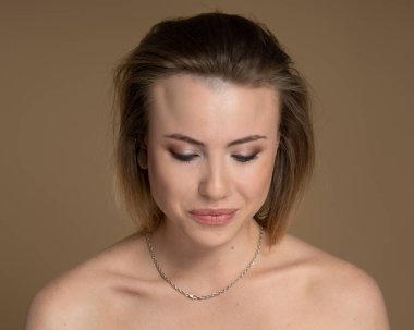 Embarrassed girl with heterochromia, nose piercing and plug in one ear, and strange hairstyle. She has amazing professional makeup and silver chain around her neck. Beige background. Studio shot