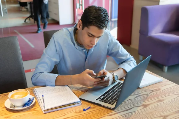若いビジネスマンが彼のスマートフォンに入力されています プロジェクトについて重要な何か 彼の隣にノートパソコンであり ホットラテのマグカップはまだテーブルの上にあります 彼はカフェに座っている — ストック写真