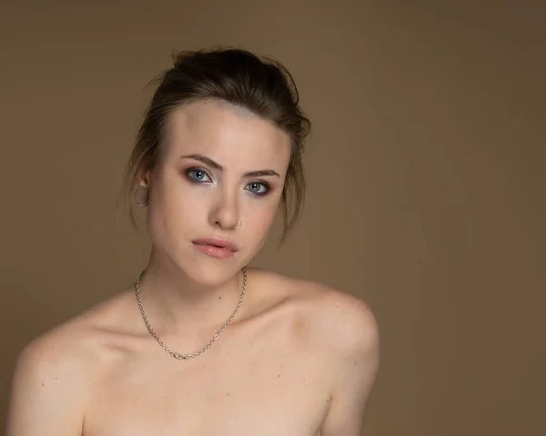 Heterokromisi olan hoş bir kız, burun piercingli ve tek kulağına takılı, ve garip bir saç stili. Boynunda inanılmaz bir profesyonel makyaj ve gümüş zincir var. Bej arka plan. Stüdyo çekimi