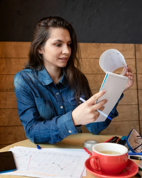 カフェに座っている青いデニムシャツの明るい女の子は ペンでメモ帳にメモを作ります — ストック写真