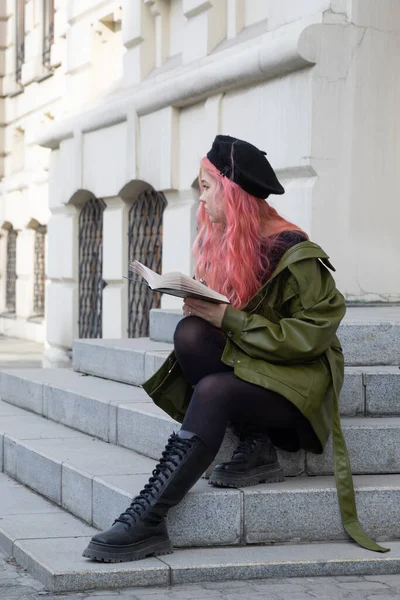 長いピンクの髪をした黒のベレー帽の賢い女の子の学生は ステップに座って本を読んで 左を見て 黒の高いブーツで 緑のジャケットで 大学の建物の背景 — ストック写真