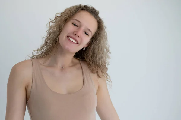 Kıvırcık saçlı, kahverengi gözlü, bej tişörtlü, gülümseyen bir kız, resim stüdyoda gri bir arka planda çekildi.