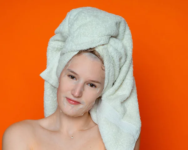 一个头发湿透 头戴白色毛巾 头戴化妆品面具 光着肩膀的女孩看上去很直率 在演播室的背景是深橙色 — 图库照片