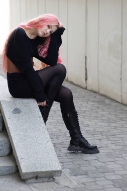 Uzun pembe saçlı kız basamaklarda tek eliyle oturuyor ve ileriye bakıyor, Basco kemeri, siyah bir kazak ve koyu renk tayt giyiyor, arkasında hoş bir bulanıklık içinde klasik bir ev var.