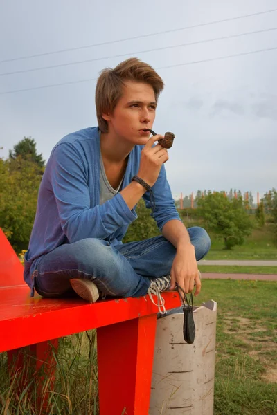 Adolescente, ragazzo seduto su una panchina e fumare una pipa — Foto Stock