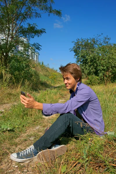 Nastoletni chłopiec siedzi na trawie i przy użyciu telefonu komórkowego — Zdjęcie stockowe