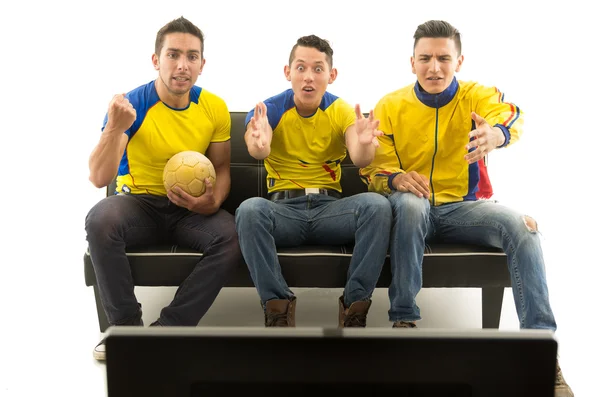 Three friends sitting on sofa wearing yellow sports shirts watching television with enthusiasm, white background, shot from behind tv — Stock Photo, Image