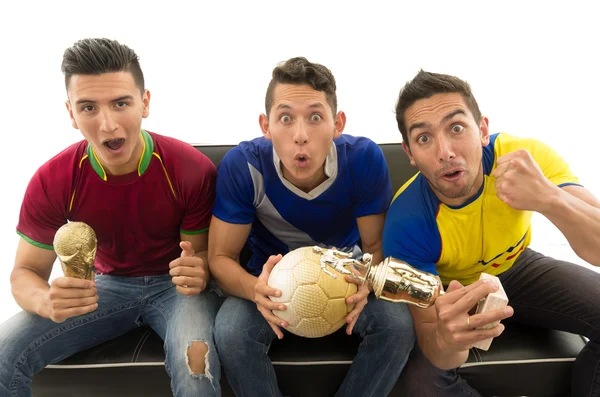 Three friends sitting on sofa wearing sports shirts, holding ball and trophy screaming cheering at camera with enthusiasm, white background, shot from above — Stock Photo, Image